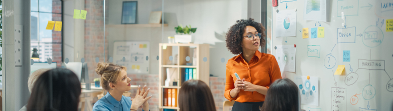A group of adults sit and stand around a table, looking at a whiteboard which is covered in colourful sticky notes and writing