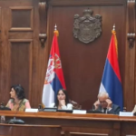 Seven people sit at a wooden bench with the Serbian flag behind them 
